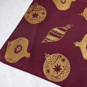 May include: A burgundy fabric table runner with a repeating pattern of gold Christmas ornaments. The ornaments are in various shapes and sizes, including round, teardrop, and star-shaped.