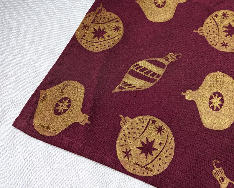May include: A burgundy fabric table runner with a repeating pattern of gold Christmas ornaments. The ornaments are in various shapes and sizes, including round, teardrop, and star-shaped.