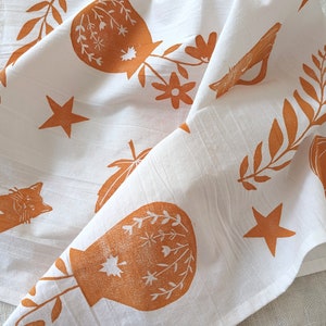May include: White cotton tea towel with a brown and orange floral print. The print features stylised leaves, flowers, stars, and a cat.