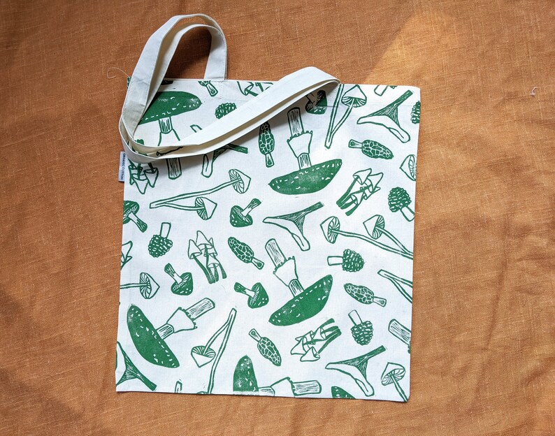 May include: A white tote bag with green mushroom illustrations. The bag has two straps and a white background.
