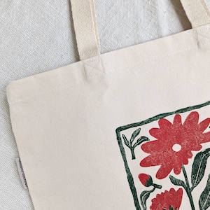 Pu&ograve; includere: Una borsa tote in tela bianca con un design floreale verde e rosso. Il design &egrave; incorniciato da un quadrato verde e presenta un grande fiore rosso con foglie verdi.