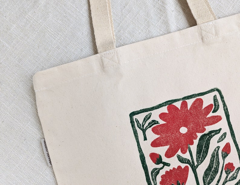 Pu&ograve; includere: Una borsa tote in tela bianca con un design floreale verde e rosso. Il design &egrave; incorniciato da un quadrato verde e presenta un grande fiore rosso con foglie verdi.