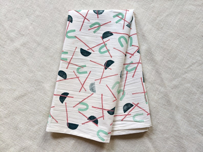 May include: A white kitchen towel with a geometric pattern of red lines, green arcs, and black semi-circles.