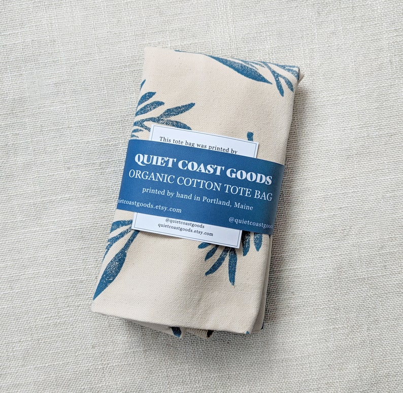 May include: A white tote bag with a blue leaf pattern. The bag is made of organic cotton and is printed by hand in Portland, Maine. The text on the bag reads "Quiet Coast Goods Organic Cotton Tote Bag".
