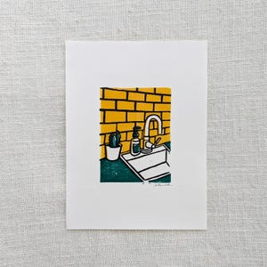 May include: A linocut print of a kitchen sink with a faucet, a small potted plant, and a bottle of soap. The sink is white and the countertop is green. The backsplash is yellow brick.