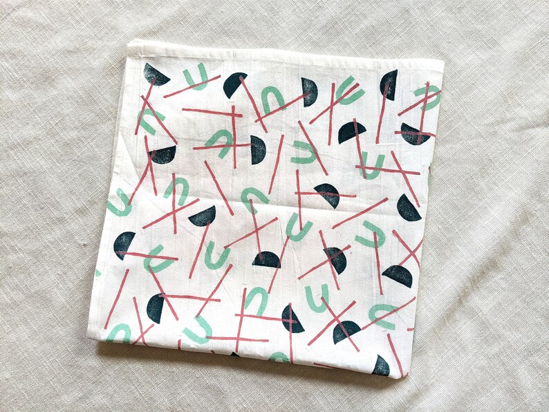May include: White cotton tea towel with a repeating pattern of black, red, and green geometric shapes. The shapes include semi-circles, lines, and U-shaped figures.
