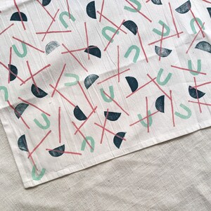 May include: A white tea towel with a repeating pattern of teal, pink, and black geometric shapes. The shapes include half circles, arches, and lines.