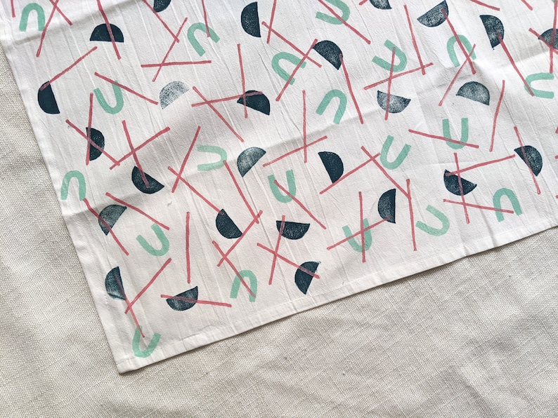 May include: A white tea towel with a repeating pattern of teal, pink, and black geometric shapes. The shapes include half circles, arches, and lines.