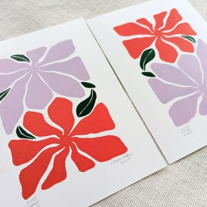 May include: Two floral prints with a purple and red flower design. The prints are on white paper and have a handwritten signature in the bottom right corner.