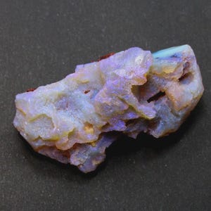 May include: A rough opal gemstone with a mix of colors, including blue, purple, yellow, and white. The stone has a textured surface and a natural, irregular shape. The opal is set against a dark background.