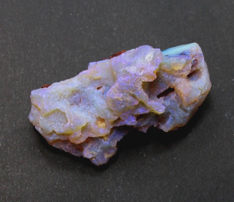 May include: A rough opal gemstone with a mix of colors, including blue, purple, yellow, and white. The stone has a textured surface and a natural, irregular shape. The opal is set against a dark background.