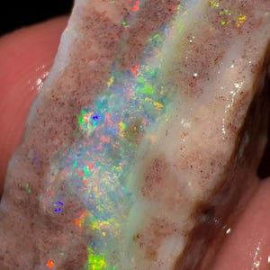 Könnte beinhalten: Nahaufnahme eines rohen Opal-Edelsteins. Der Stein hat eine hellbraune Matrix mit Bereichen von schillernden Farben, darunter Blau, Grün und Orange. Der Opal wird in der Hand einer Person gehalten, mit dunklem Hintergrund.