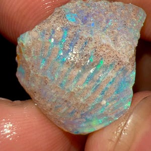 Könnte beinhalten: Ein roher, unpolierter Opal-Edelstein mit einer texturierten, beigefarbenen Oberfläche und markanten parallelen Rillen. Der Stein zeigt lebhafte Blitze aus irisierendem Blau, Grün und Violett über seine unregelmäßige, zerklüftete Form.
