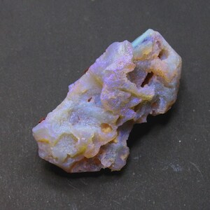 May include: A rough, natural opal gemstone with a mix of colors. The stone has a light blue base with areas of yellow, purple, and orange. The opal has a textured, uneven surface.