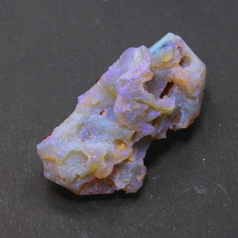 May include: A rough, natural opal gemstone with a mix of colors. The stone has a light blue base with areas of yellow, purple, and orange. The opal has a textured, uneven surface.