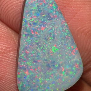 May include: A teardrop-shaped opal gemstone featuring a vibrant play-of-color with scattered flecks of bright red, electric blue, and neon green against a translucent, milky white base, polished to a smooth, rounded finish.
