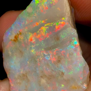 May include: A raw opal gemstone with a milky white base. The stone exhibits iridescent flashes of colour, including red, orange, yellow, green, and blue. The opal is held in a hand, showing its natural, unpolished state.