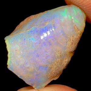 以下が含まれることがあります： 虹色の青、緑、紫が特徴の、色の遊びが見られるラフオパール原石。 石は質感のある薄茶色のマトリックスを持ち、黒い背景に対して指で持たれています。