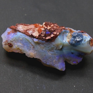 May include: A rough-cut opal gemstone with a mix of colors. The stone features a base of pale blue and white, with flashes of purple and blue. The top of the stone has a reddish-brown, textured matrix.
