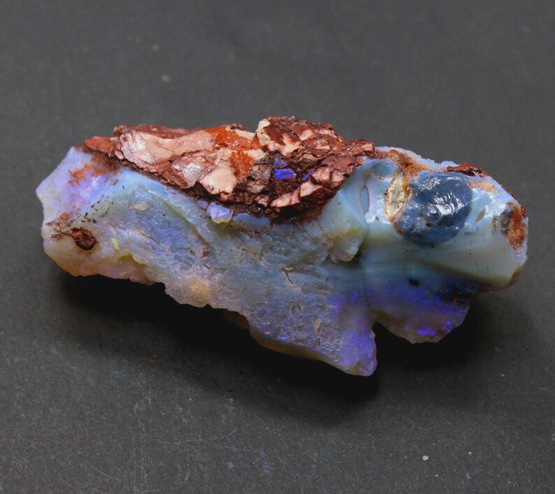 May include: A rough-cut opal gemstone with a mix of colors. The stone features a base of pale blue and white, with flashes of purple and blue. The top of the stone has a reddish-brown, textured matrix.