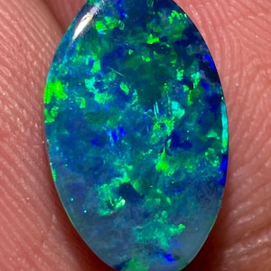 May include: An oval-shaped loose gemstone featuring a deep blue base color with vibrant, scattered flashes of bright green and electric blue play-of-color throughout the surface, displaying a polished, smooth, and reflective finish.