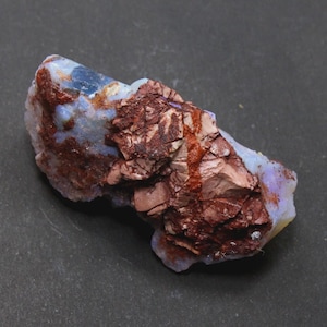May include: A raw, uncut opal specimen with a mix of colors. The stone features a base of pale blue and white, with areas of deep reddish-brown and a rough, textured surface. The opal is displayed against a dark background.