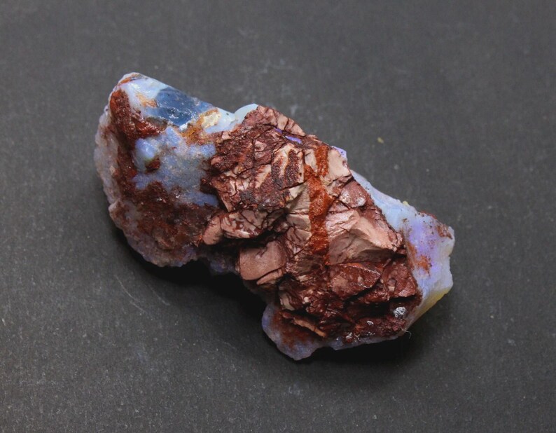 May include: A raw, uncut opal specimen with a mix of colors. The stone features a base of pale blue and white, with areas of deep reddish-brown and a rough, textured surface. The opal is displayed against a dark background.