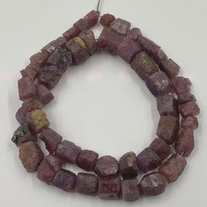 May include: Strand of raw ruby beads. The beads are a deep reddish-purple color with a rough, irregular texture. The beads are strung on a thin, silver-colored wire. The beads are of varying sizes and shapes, with a few showing hints of yellow.