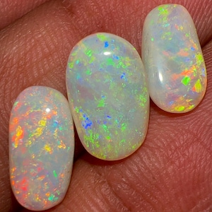 May include: Three oval-shaped opals with a milky white base and vibrant flashes of color. The opals display a play-of-color effect, with hues of green, blue, orange, and red. The gemstones are smooth and polished, showcasing their natural beauty.