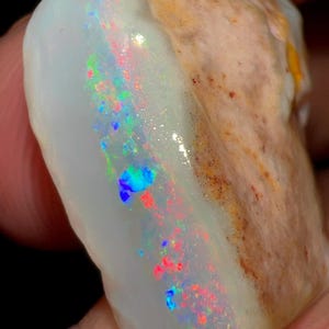 May include: A raw opal gemstone featuring a milky white base with a prominent vertical band of iridescent play-of-colour, displaying vibrant flashes of neon green, electric blue, and bright red against a textured, tan-coloured rocky matrix.