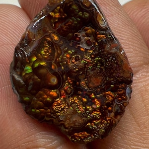 May include: A polished fire agate gemstone featuring a dark brown base with iridescent flashes of green, orange, and gold. The surface displays a botryoidal, bubbly texture with distinct color play throughout the stone's irregular, rounded shape.