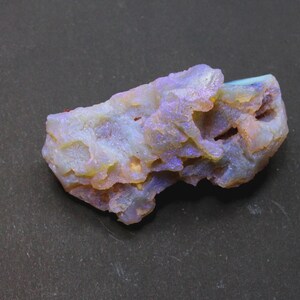 May include: A rough, uncut opal gemstone with a mix of colors. The stone has a base of white and yellow, with patches of blue and purple. The surface is textured and irregular, with small crevices and variations in color. The opal is set against a dark gray background.