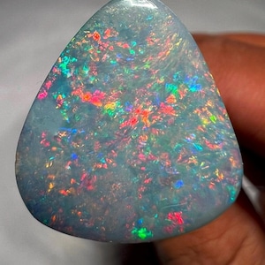 May include: A polished, triangular opal gemstone with a pale blue base color. The stone displays a vibrant play-of-color, with flashes of red, green, yellow, and blue throughout its surface. The opal is held in a hand, showcasing its natural beauty.