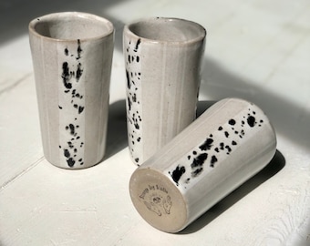 May include: Three white ceramic mugs with black splatter paint and a white stripe down the side. The mugs are on a white wooden surface. The bottom of one mug has the text "Sleepy Joy Studio" printed on it.