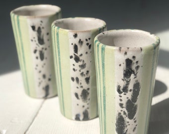 May include: Three ceramic cups with a green and white striped pattern and black splatter paint. The cups are lined up on a white wooden surface.