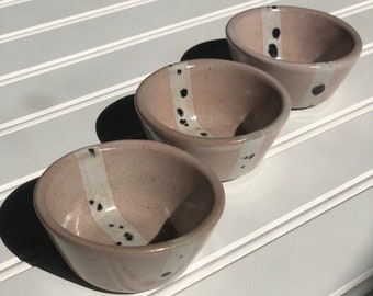 May include: Three small ceramic bowls with a pink glaze and black and white speckled design. The bowls are arranged in a row on a white surface.