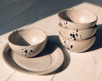 May include: A set of four white ceramic bowls with black speckles. The bowls are stacked on top of each other. There is a matching white ceramic plate with a geometric pattern.