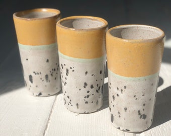May include: Three ceramic shot glasses with a speckled white glaze and a yellow rim. The glasses have a thin band of light green glaze around the middle.