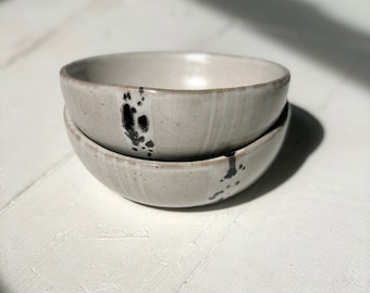 May include: Two white ceramic bowls with black speckles stacked on top of each other. The bowls are slightly rounded and have a smooth, glossy finish.