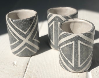 May include: Three gray ceramic cups with a white geometric pattern. The cups are on a white wooden surface.