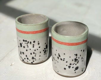 May include: Two ceramic mugs with a speckled white glaze and black speckles. The mugs have a light green band with a thin orange band around the top.