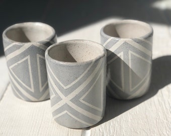 May include: Three gray ceramic cups with a white geometric pattern. The cups are arranged in a triangular shape on a white wooden surface.
