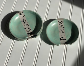 May include: Two teal ceramic dishes with a white and black speckled stripe design. The dishes have a rounded shape and a slight lip around the edge.