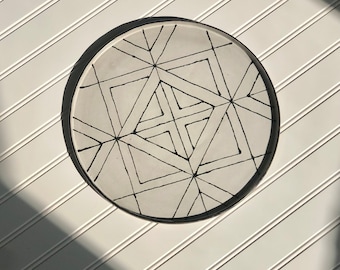May include: A white ceramic plate with a black geometric design. The design features a series of intersecting lines and squares.