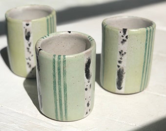 May include: Three green ceramic cups with black and white stripes. The cups are on a white surface.