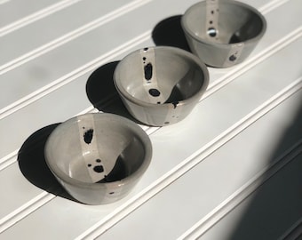 May include: Three small grey ceramic bowls with black splatter paint and a white stripe. The bowls are arranged in a row on a white surface.