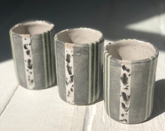 May include: Three ceramic mugs with a striped pattern in shades of gray, black, and white. The mugs have a speckled glaze and a slightly textured surface.