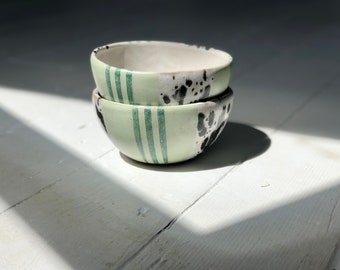 May include: Two small ceramic bowls stacked on top of each other. The bowls are green with black and white speckles and green stripes.