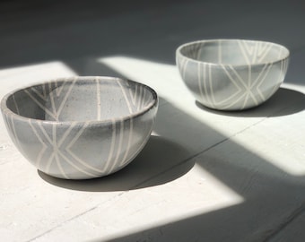 May include: Two small grey ceramic bowls with a white geometric pattern. The bowls are sitting on a white surface.