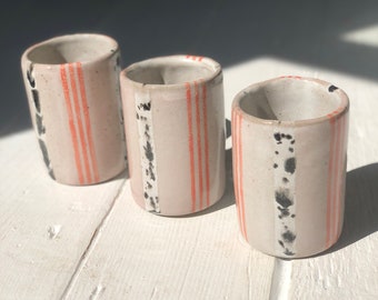 May include: Three small ceramic cups with a striped pattern in shades of gray, white, and orange. The cups are lined up on a white wooden surface.